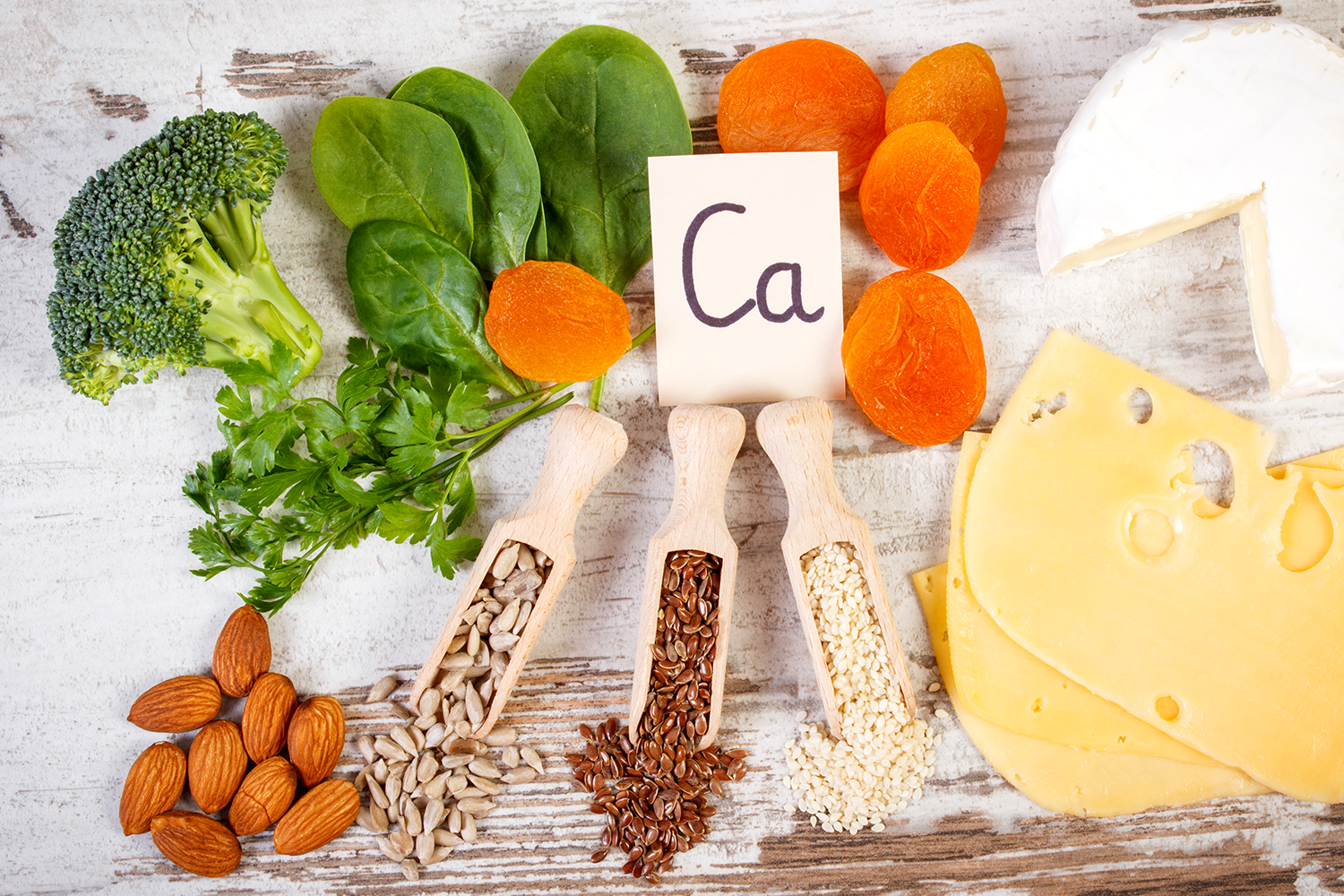 Broccoli, apricots, cheese, seeds, nuts, and parsley lying upon a wood surface surrounding a card reading "Ca" for a blog post about Hypercalcemia